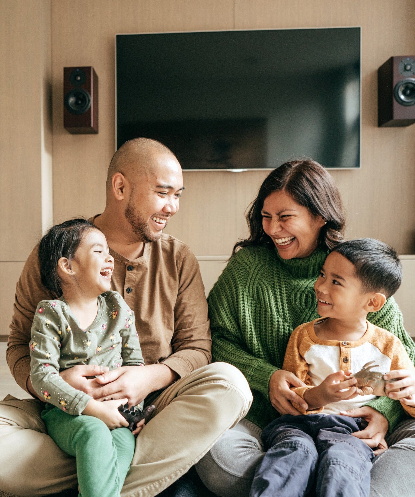 Parent holding two children on a couch with a tv in the background.